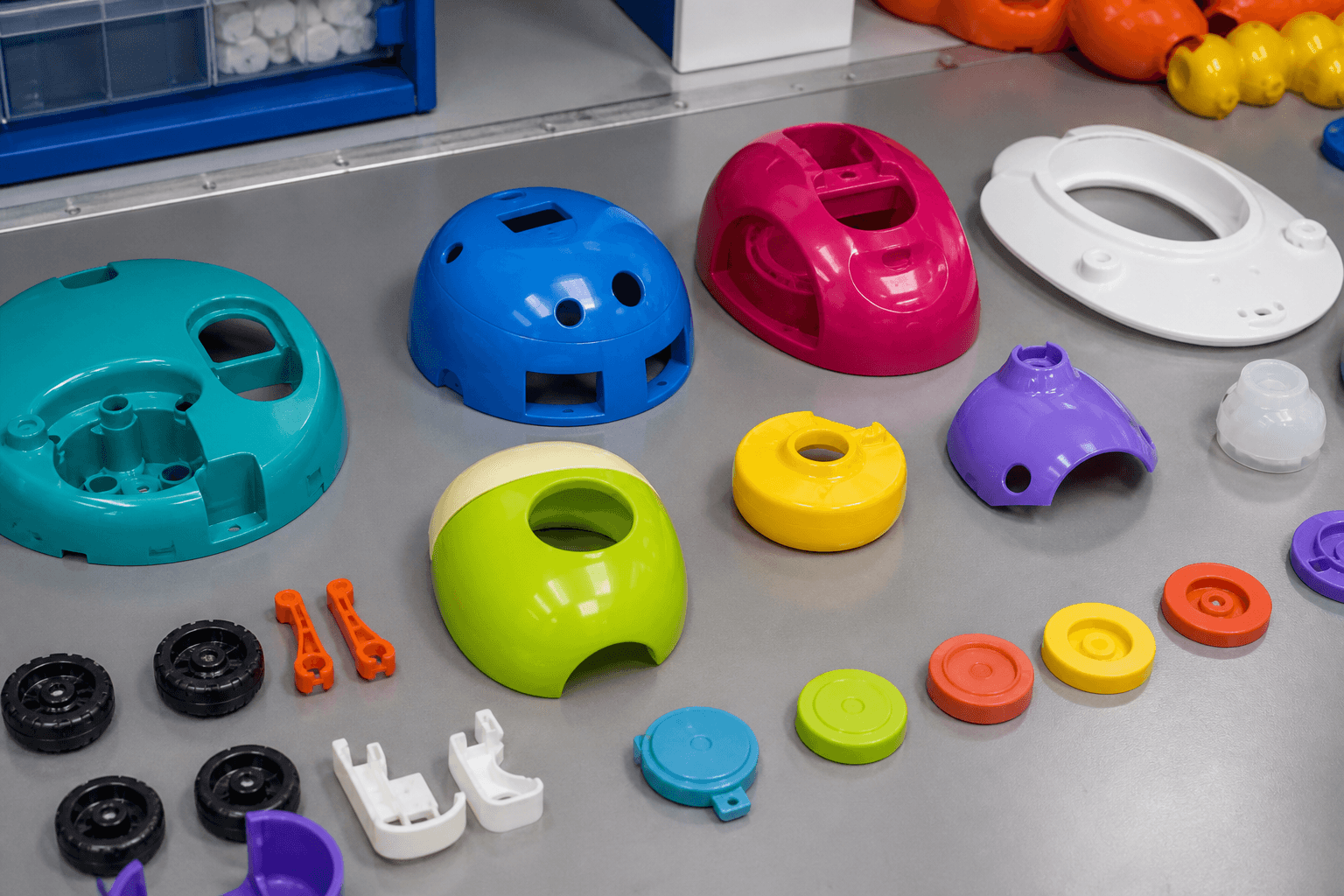 Brightly colored molded toy shells and domes in blue, pink, teal, green, and purple, with small wheels, caps, and white brackets on a steel table